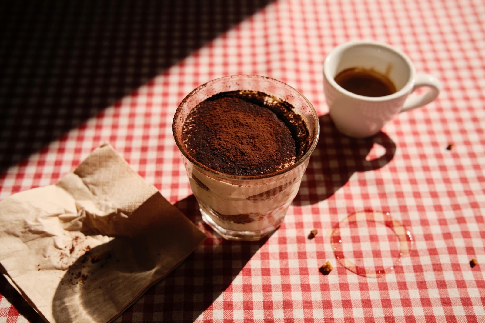 Un tiramisu au cacao dans un verre, une tasse d'expresso et une serviette en papier froissée sur la nappe à carreaux.